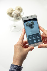Woman photobraph on her phone various of round raw organic sweets with cacao chocolate and sesame. Homemade cooking