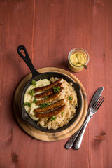 Kleine Bayrische Nürnberger Rostbratwürste mit Sauerkraut, Kartoffelpüree und Senf in rustikaler Gusseisenpfanne auf Holz Brett