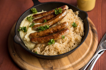 Kleine Bayrische Nürnberger Rostbratwürste mit Sauerkraut, Kartoffelpüree und Senf in rustikaler Gusseisenpfanne auf Holz Brett