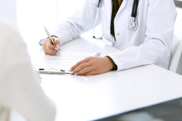 Woman doctor and patient sitting and talking at medical examination at hospital office, close-up. Therapist filling up medication history records. Medicine and healthcare concept