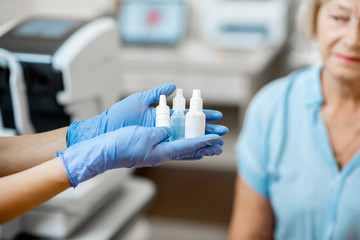 Nurse in medical gloves holding white bottles or droppers with eye medicine with senior patient on...