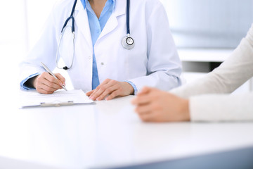 Woman doctor and patient sitting and talking at medical examination at hospital office, close-up. Physician filling up medication history records. Medicine and healthcare concept