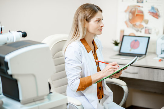 Beautiful female ophthalmologist writing a medical recipe or patient's history while sitting in ophthalmologic office