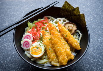 Close up of bowl of gourmet Japanese udon noodle soup with colorful toppings of pickled ginger, boiled egg, swirly pattern narutomaki, and breaded prawns