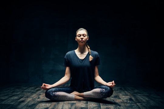 Young attractive woman practicing yoga sitting in lotus pose and meditating in dark room