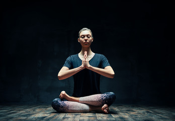 Young sporty woman practicing yoga sitting in lotus pose with namaste in dark room