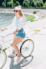 Naklejka na ściany i meble smiling blonde beautiful girl riding bike near river in summer