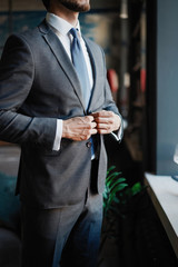 Stylish young bearded man in a suit is standing near the window and looking far