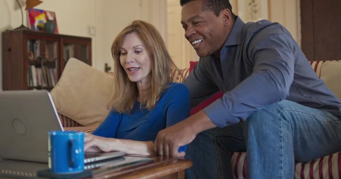 Older African American and Caucasian husband and wife using laptop computer together in living room. Mature homeowner married couple smiling while planning trip online. Slow motion 4k handheld