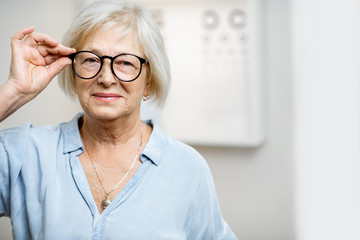 Portrait of a happy senior woman wearing eyeglasses in front of eye chart in ophthalmology office. Concept of checking eyesight and selecting glasses in older age