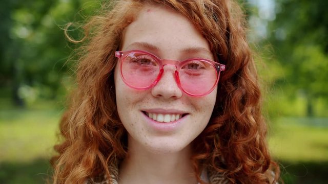 Slow motion of attractive teenager with red curly hair wearing sunglasses smiling in green park alone looking at camera. Youth and positive emotions concept.