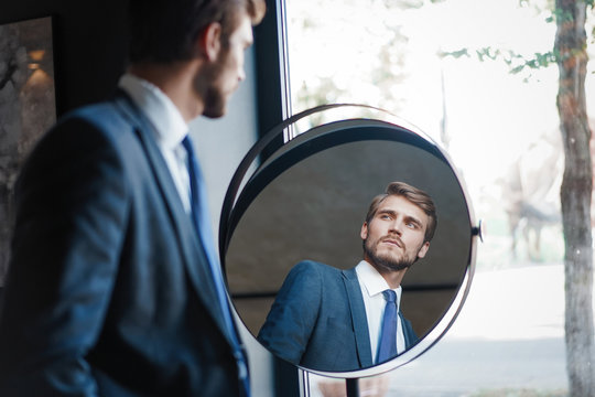 Reflection of handsome young man in full suit standing near the window in front of the mirror indoors