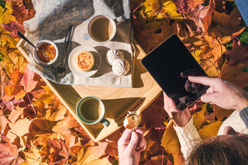 Couple of young people are looking on a tablet having morning breakfast on a wooden tray in the autumn park with colorful maple leaves. Aerial view