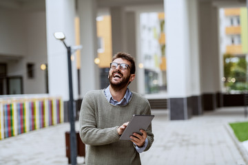 Laughing handsome bearded Caucasian fashionable man in gray sweater and with eyeglasses standing outdoors and using tablet.