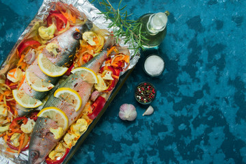  Baked fish with vegetables and lemon, tomatoes and spices in a baking dish. Top view on a blue background. Copy space.