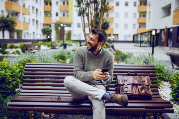 laughing handsome stylish businessman typing message on smart phone while sitting on the bench in...