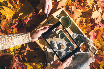 Couple of young people are taking a picture on a tablet of morning breakfast on a wooden tray in the autumn park with colorful maple leaves. Aerial view