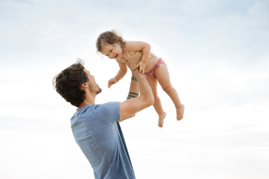 Low angle view of father lifting laughing daughter against sky at beach