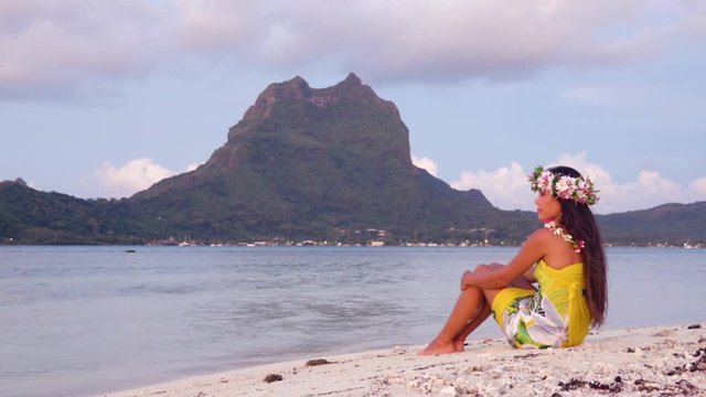 French Polynesia Beach Vacation Travel Holidays. Woman relaxing sitting down on paradise beach on motu, Bora Bora. Girl wearing traditional pareo and flower head crown in Tahiti French Polynesia