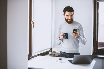 Portrait of happy man with beard standing in kitchen and using his smartphone while drink coffee