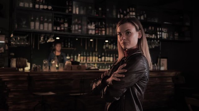 beautiful young woman in a trendy bar folds her arms over her chest, bartender in background is preparing a cocktail