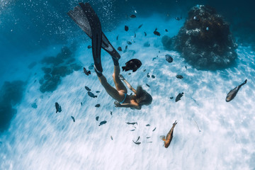 Freediver girl with fins glides over sandy bottom with fishes in transparent sea