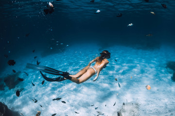 Freediver girl with fins glides over sandy bottom with fishes in transparent sea