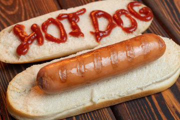 Focus on the sausage. Hot dog bun with fried sausage on dark wooden background. On the bun is written in red ketchup - hot dog