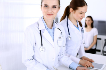 Fototapeta premium Group of doctors at work in hospital with patient in a queue at the background. Physician filling up medical documents or prescription while standing at reception desk. Data in medicine and health