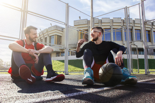 Two friends are sitting on a basketball court in a sports uniform, talking and laughing. Half time in the middle of a basketball game. Friends communicate and enjoy. Two friends with a basket ball.
