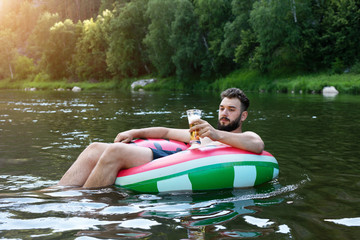 Hipster with inflatable circle on summer nature background. young man with a beard swims in a life buoy and holds a glass with beer