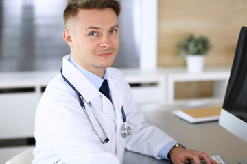 Doctor man sitting behind a computer at workplace in clinic or hospital office. Medic headshot and medicine concept