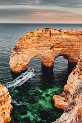 Foto auf Acrylglas Strand Marinha, Algarve, Portugal Wunderschönes kristallklares Meerwasser und ein Boot auf der Vorderseite ein Naturküsten-Felsbogen bei farbenfrohem Sonnenuntergang. Praia da Marinha, berühmter Strand, Küste der Algarve, Lagoa, Portimao in Südportugal, Atlantik