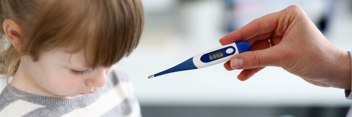 Obraz premium Portrait of thoughtful girl sitting in modern office of professional physician and looking down. Caring woman pediatrist holding thermometer. Clinic examination concept