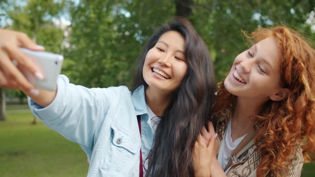 Cute girls friends are taking selfie with smartphone camera posing in green park hugging smiling having fun. Modern gadgets, people and friendship concept.