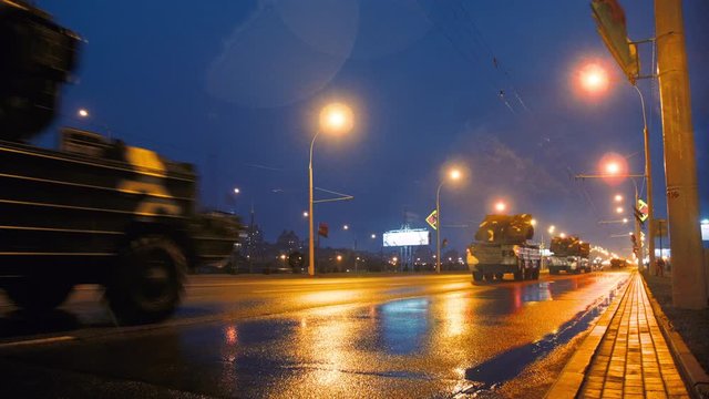 A convoy of military equipment rides through the city at night with headlights on