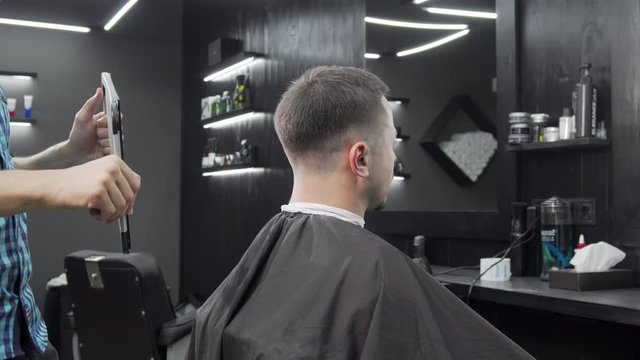 Barber holding a mirror for his male client to check out new haircut. Man examining his new hairstyle in the mirror at local barbershop. Profession, service, hair concept