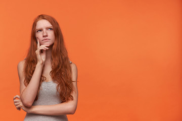 Cute young pensive female with wavy foxy hair lond hair looking upwards while thinking, isolated over orange background, wearing casual grey shirt, holding hand on her face