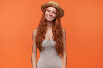 Horizontal portrait of pleasant looking happy young female with wavy foxy hair isolated over orange background, wearing casual clothes and straw hat, positive emotions concept
