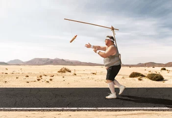 Fotobehang Funny overweight man chasing the hot dog on the stick through the empty road with copy space © rangizzz