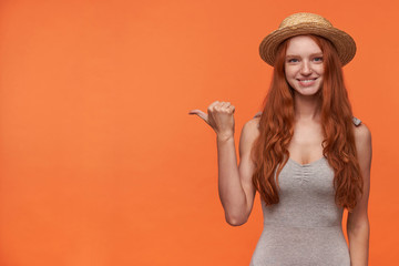 Attractive cheerful young lady with wavy foxy hair looking to camera positively and showing with thumb aside, posing over orange background in casual clothes