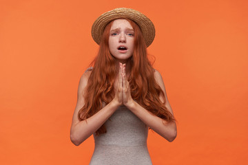 Upset young pretty woman with wavy red lond hair standing over orange background, keeping palms together in praying gesture, hoping for better, wearing casual clothes