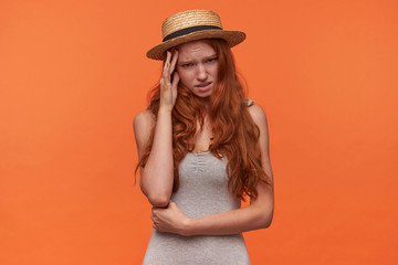 Studio shot of beautiful young redhead female with wavy foxy hair hiding behind her palm, looking guilty to camera with ashamed face, isolated over orange background