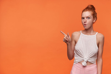 Horizontal shot of pretty young redhead woman with bun hairstyle wearing casual clothes, looking to camera and moving eyebrows, pointing away with forefinger, isolated over orange background
