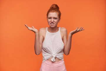 Horizontal indoor photo of pretty young woman with foxy hair combed in bun posing over orange background, shrugging perplexedly with raised hands and frowning her face
