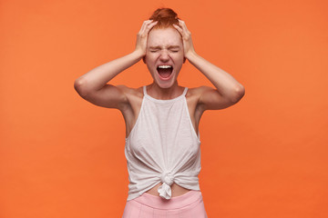 Studio close-up of stressed young woman with foxy hair combed in bun standin over orange background, keeping hands on temples and screaming loud with closed eyes