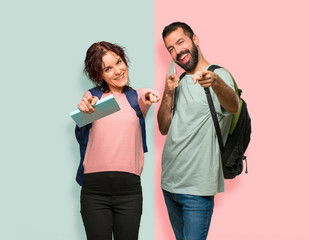 Two students with backpacks and books points finger at you while smiling on colorful wall