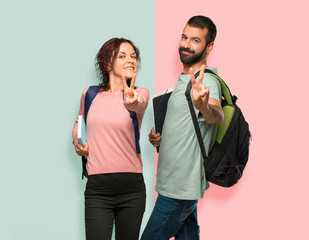 Two students with backpacks and books smiling and showing victory sign on colorful wall