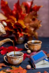 Morning coffee in the mugs and the coffee pot pours coffee in a mug on a dark wooden background with maple leaves and and a book