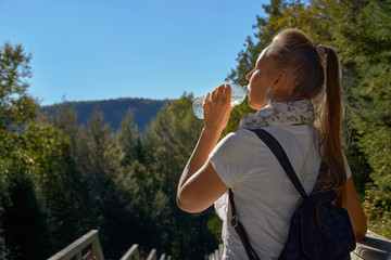Naklejka premium Attractive young blonde in a white T-shirt drinking water from a bottle, standing in the mountains on a sunny summer day. Retouching, bright colors